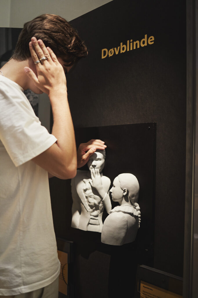 A person in a white t-shirt holds his hand to his ear while touching a stone sculpture