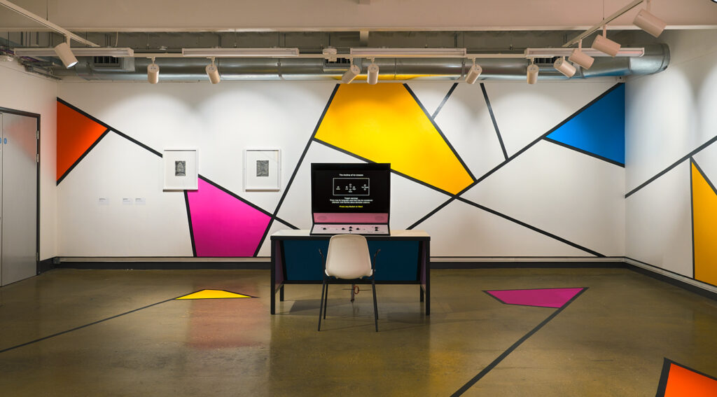 A computer screen in a room decorated with geomtric shapes in bright pink, blue and yellow colours