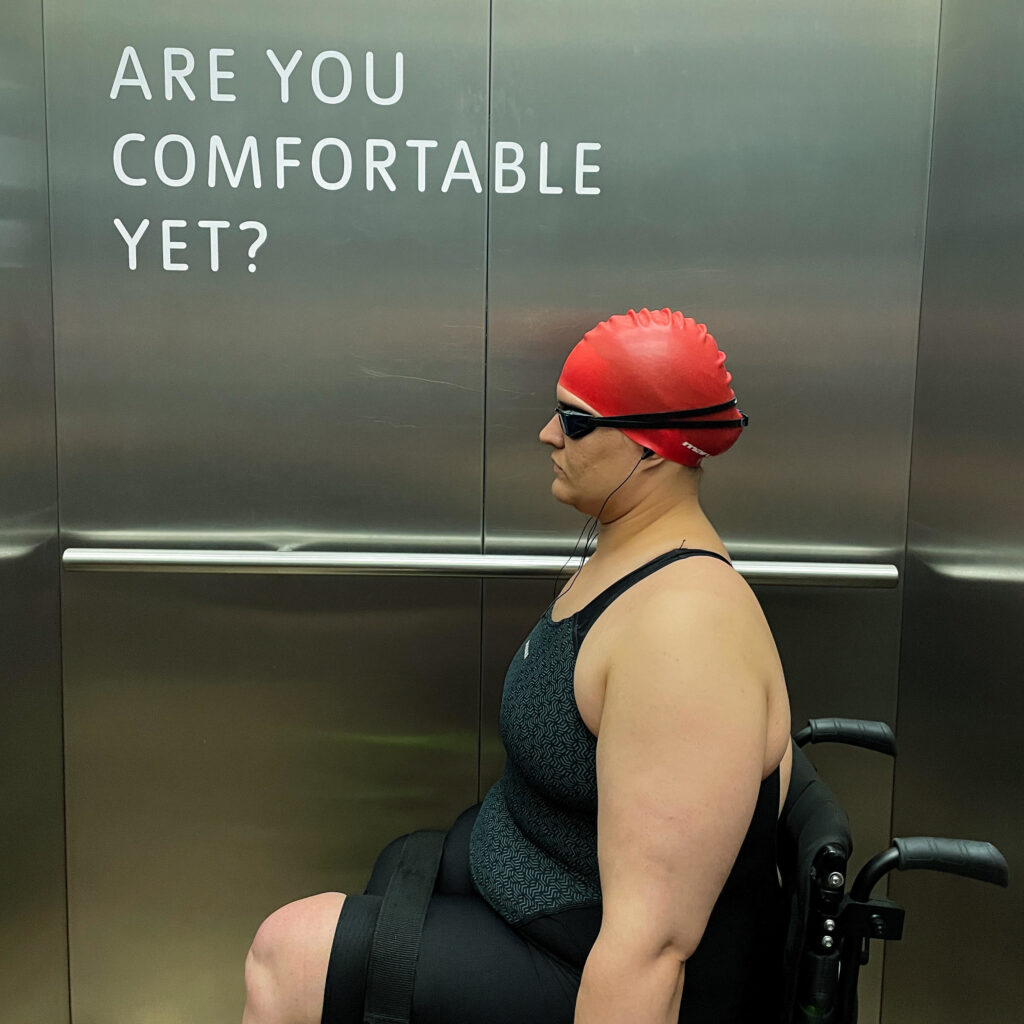 A woman in a wheelchair wearing a swimming hat and goggles. She sits in a lift next to a sign that says Are you comfortable yet?