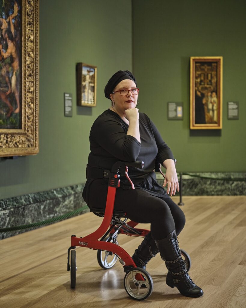 Stacey Smith is a young white woman. She is pictured sitting on her red walker, and wears a black outfit, glasses and a black headscarf