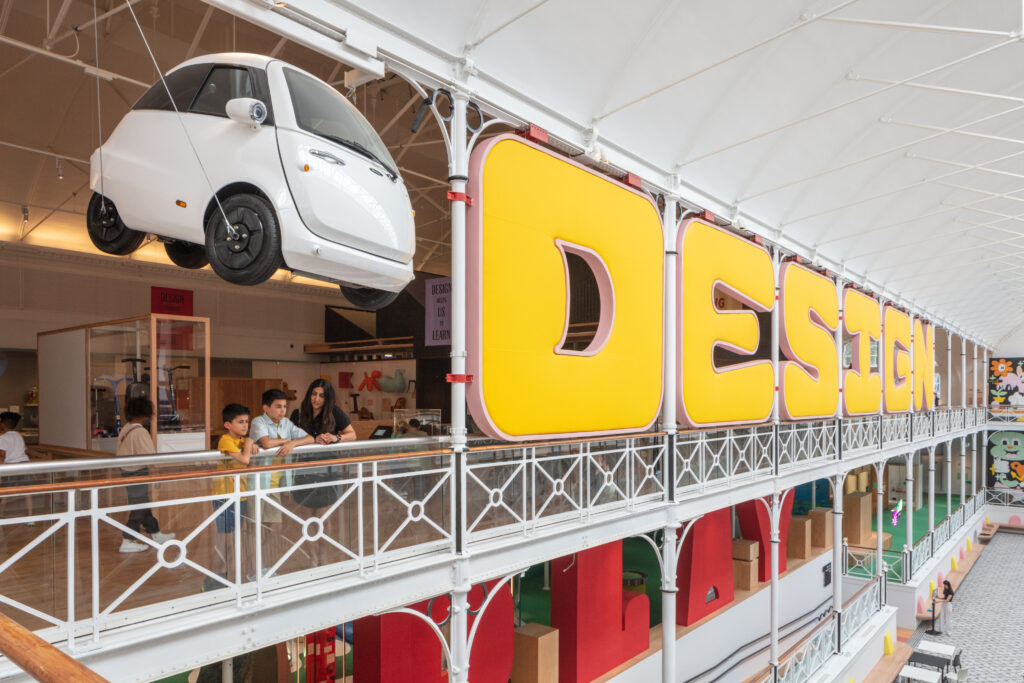 Large museum atrium with a white car hanging from roof and the word 'Design' in brig friendly yellow letters 