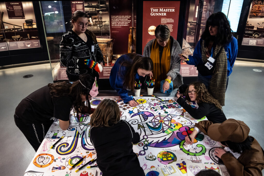 A group of people gather around a colorful table covered in drawings and markers, collaborating on an art project in a museum exhibit space. Some are coloring while others watch and discuss.