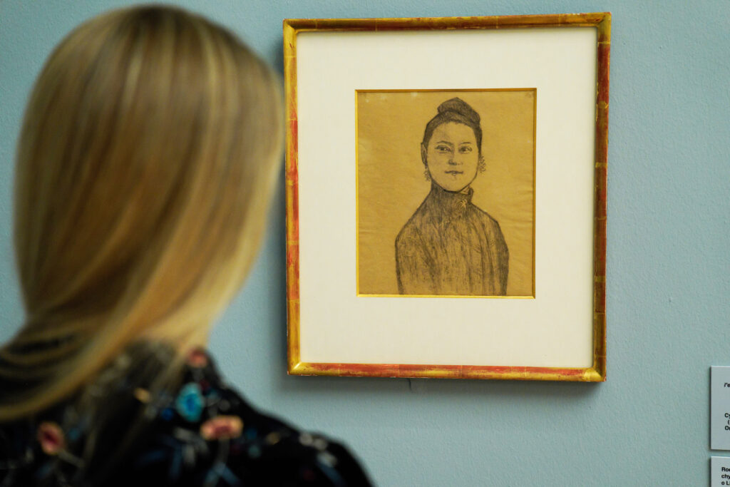 A person with long blond hair views a framed pencil portrait of a woman, displayed on a blue wall in an art gallery.